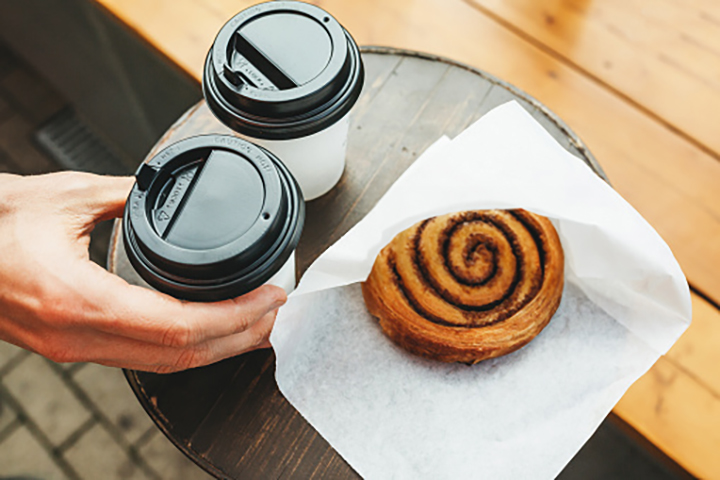 Two to go coffees and a cinnamon roll on a table with a hand grabbing one of the coffees.